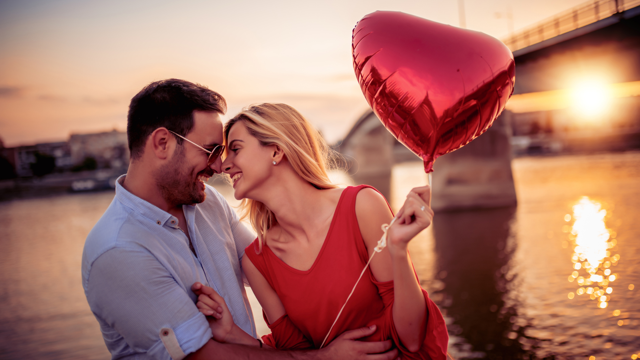 image d'un couple se regardant le sourire aux lèvres le tout avec un ballon en forme de coeur à la main devant un fleuve pendant un coucher de soelil